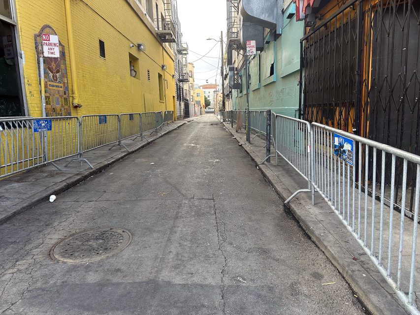 A narrow urban alleyway lined with yellow and green buildings, metal barricades on both sides, and “No Parking” signs posted on the walls.