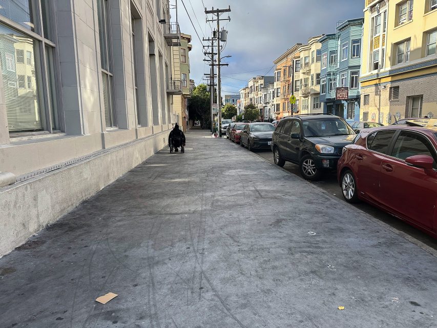 A wide city sidewalk with a person pushing a stroller, cars parked along the street, and multi-story buildings on both sides.