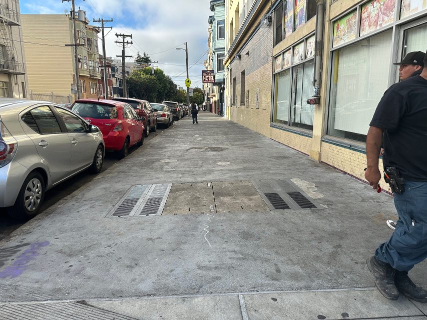 A sidewalk in an urban area with parked cars on the left, buildings on the right, and a few people walking in the distance. A man stands on the right edge of the image.
