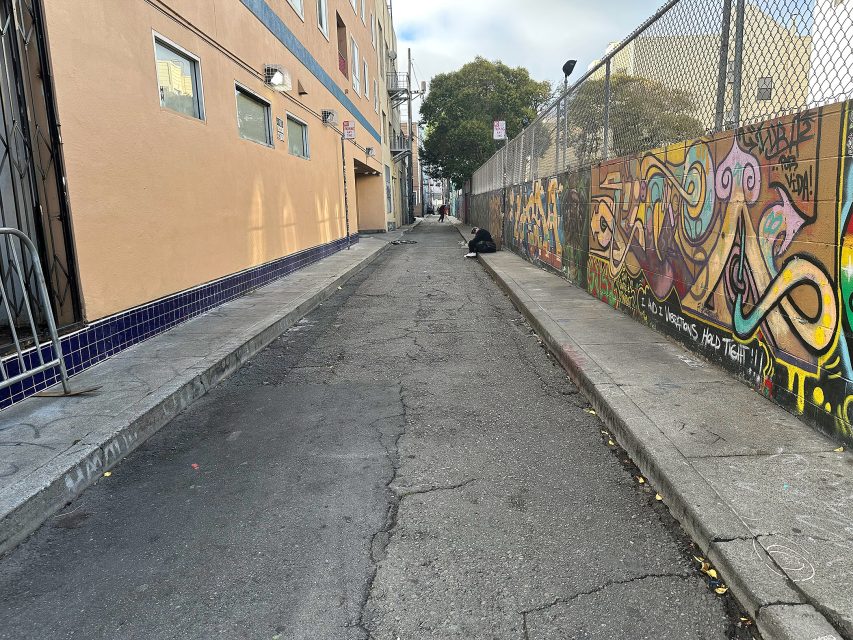 Narrow urban alley with cracked pavement, graffiti-covered wall on the right, and a person sitting near the end of the alley.