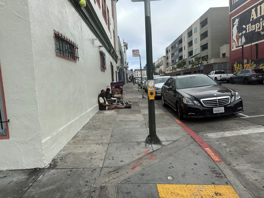 Two people sit on the sidewalk near a building, with a black car parked on the street and apartments visible in the background.
