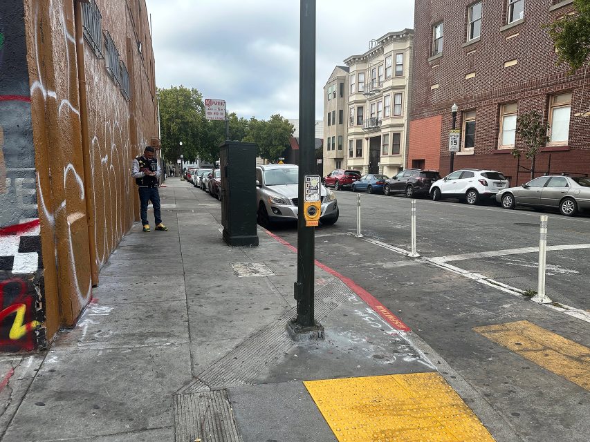 A city street corner with a crosswalk button on a pole, parked cars, graffiti on a building, and a pedestrian walking on the sidewalk.