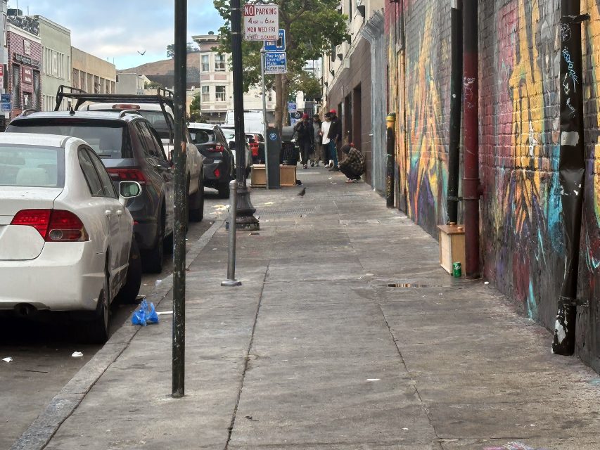 Urban sidewalk with parked cars, graffiti-covered wall, scattered litter, and several people gathered in the distance.