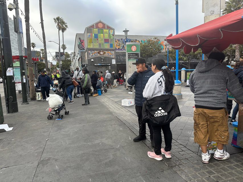 A group of people gather and stand around a busy urban sidewalk, some with carts and bags, near a colorful mural and vendor stands under a cloudy sky.