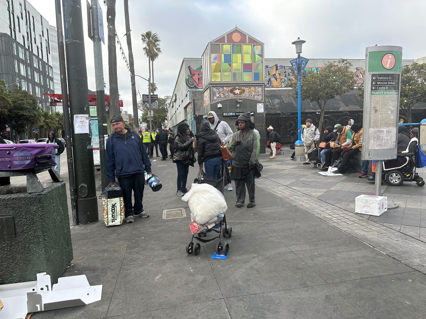 People stand and sit in line on a city sidewalk near a colorful building, some with bags and carts, in an urban outdoor setting.