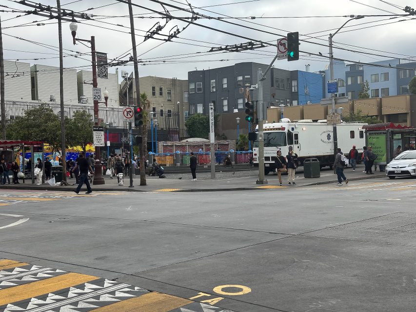 A busy city intersection with pedestrians, buildings, power lines, and a large white vehicle parked near the corner under a cloudy sky.