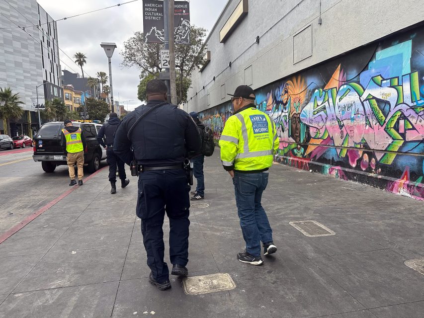 Police officers and city workers walk along a sidewalk lined with colorful graffiti murals in an urban area.
