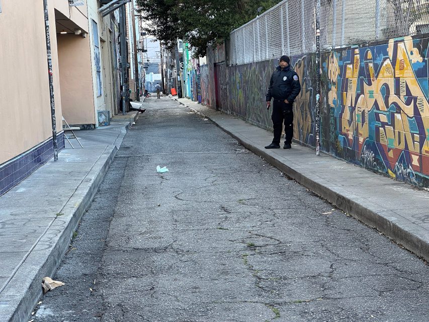 A police officer stands on the sidewalk of a graffiti-covered alley, looking down the empty street lined with buildings and a chain-link fence.