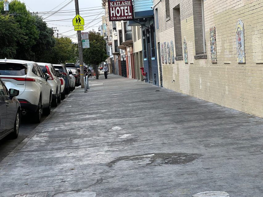 A sidewalk lined with parked cars next to a beige brick building; a sign for Katlash Hotel hangs above the street.