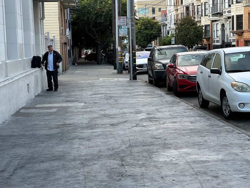A man stands on a city sidewalk next to a white building, with parked cars lined up along the street and apartment buildings in the background.