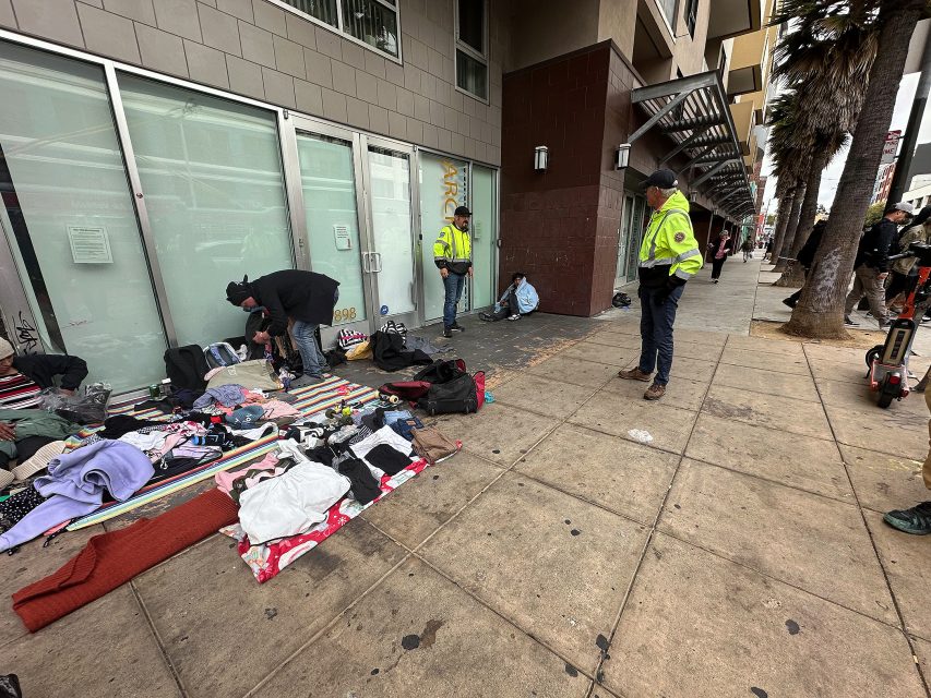 Three people, including two in high-visibility jackets, stand on a city sidewalk near laid-out clothes and belongings in front of a building with glass windows.
