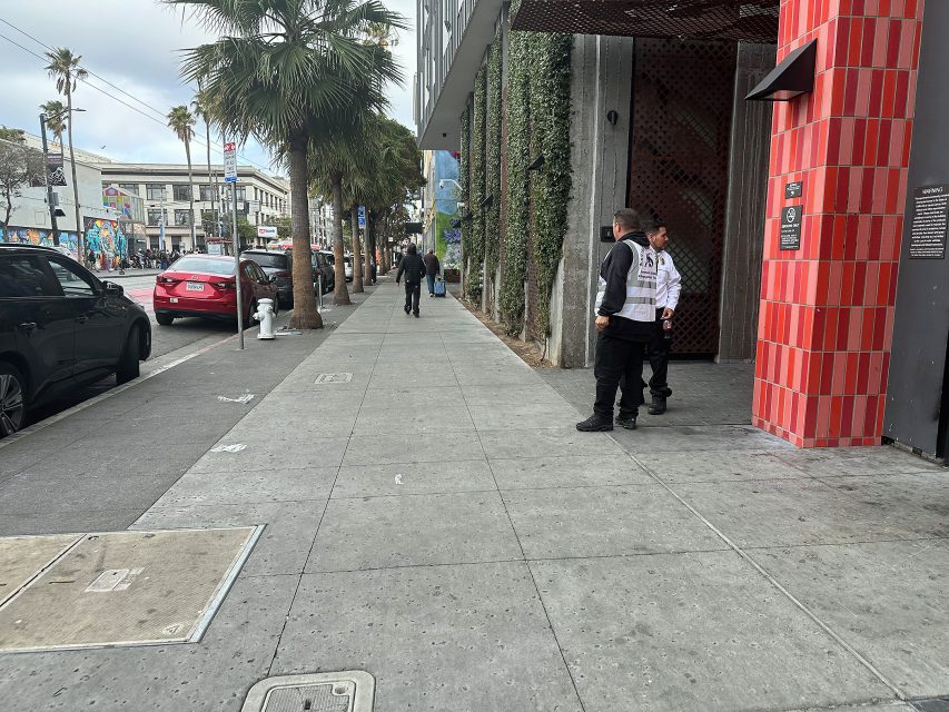 Two security guards stand near a building with red tiles on a city sidewalk; cars are parked along the street and palm trees line the road.