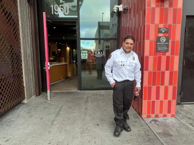 A security guard stands in front of a building entrance labeled "1950" next to a red-tiled wall with a "No Smoking" sign. He is holding a drink bottle.