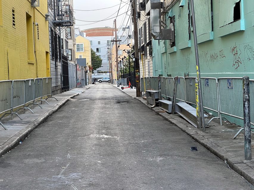 A deserted urban alleyway lined with metal barricades, adjacent to yellow and green buildings with graffiti and utility wires overhead.