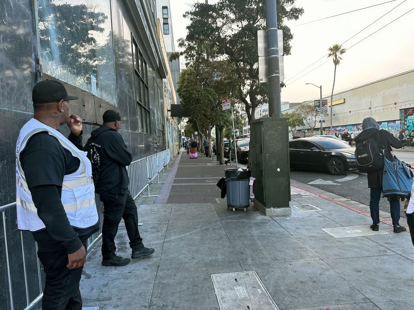Two men in black clothing stand on a city sidewalk near a building, while other pedestrians walk past and cars are parked along the street.