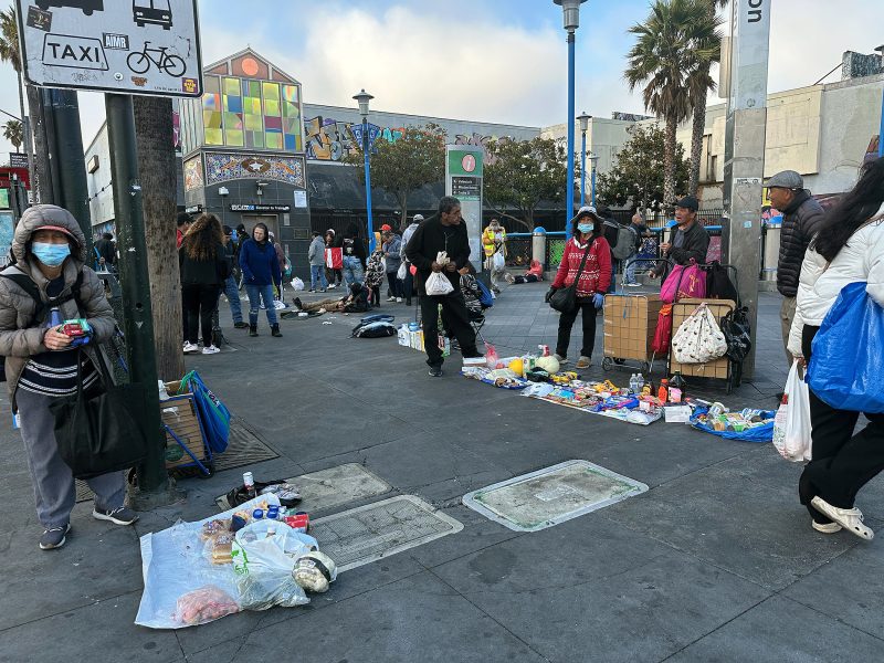 People sell various goods laid out on the sidewalk in an urban outdoor market setting; some wear masks, and a colorful building is visible in the background.