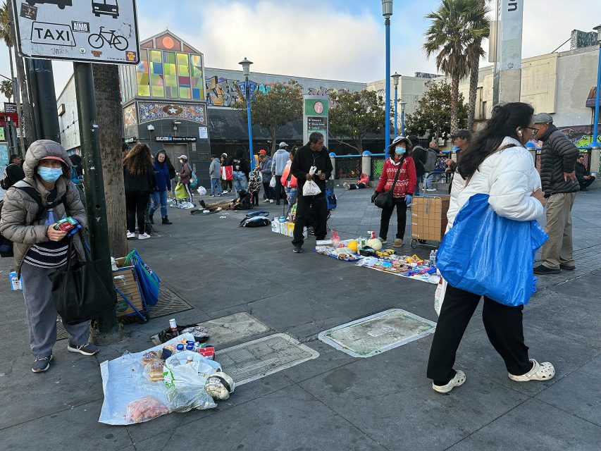 People walk by street vendors selling various goods laid out on the ground in an urban plaza; some vendors and shoppers are wearing masks.