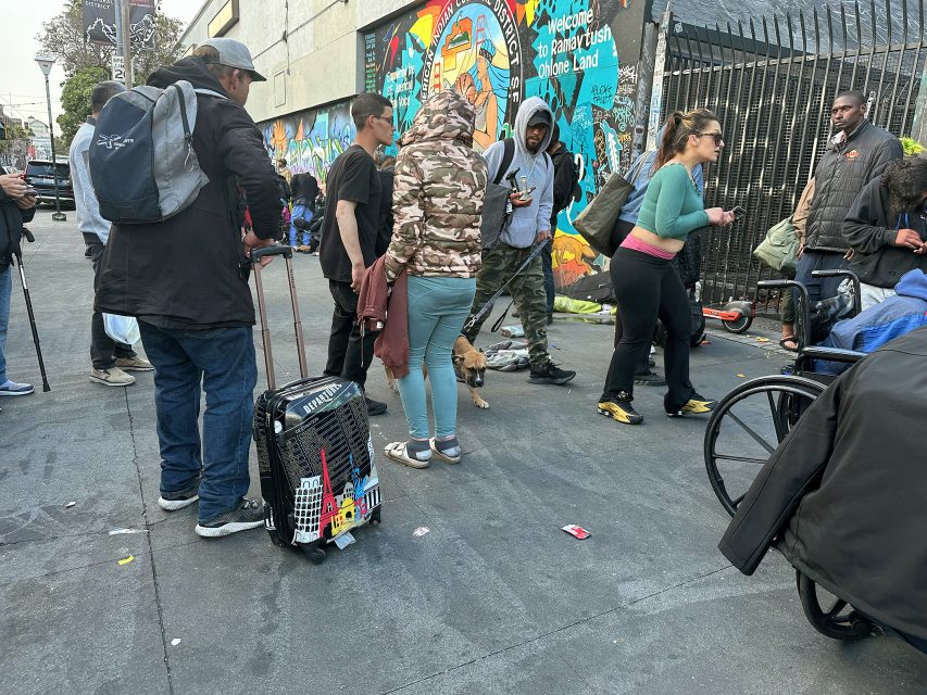 A group of people, some with luggage and backpacks, stand and walk on a city sidewalk near a colorful mural; one person is in a wheelchair and a dog is present.