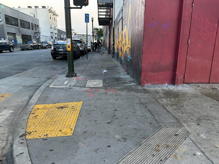Urban sidewalk with a textured yellow ramp and tactile paving, bordered by a red building with graffiti; cars are parked along the street and pedestrians walk in the distance.