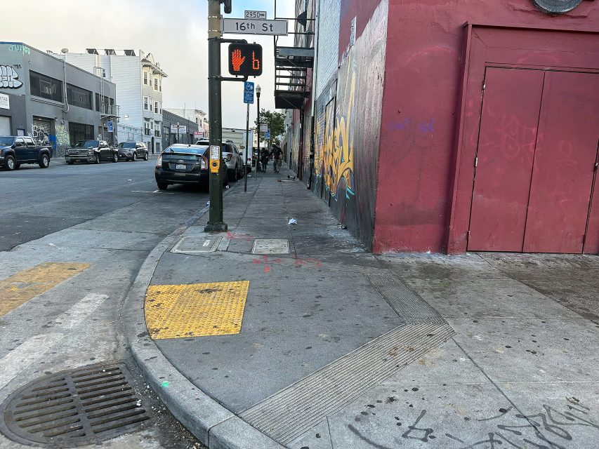 A city sidewalk near a red building with graffiti, a street crossing signal, and some parked cars along the road.