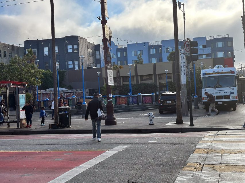 A man walks a small white dog across an urban street near a bus stop, with people and vehicles in the background on a cloudy day.
