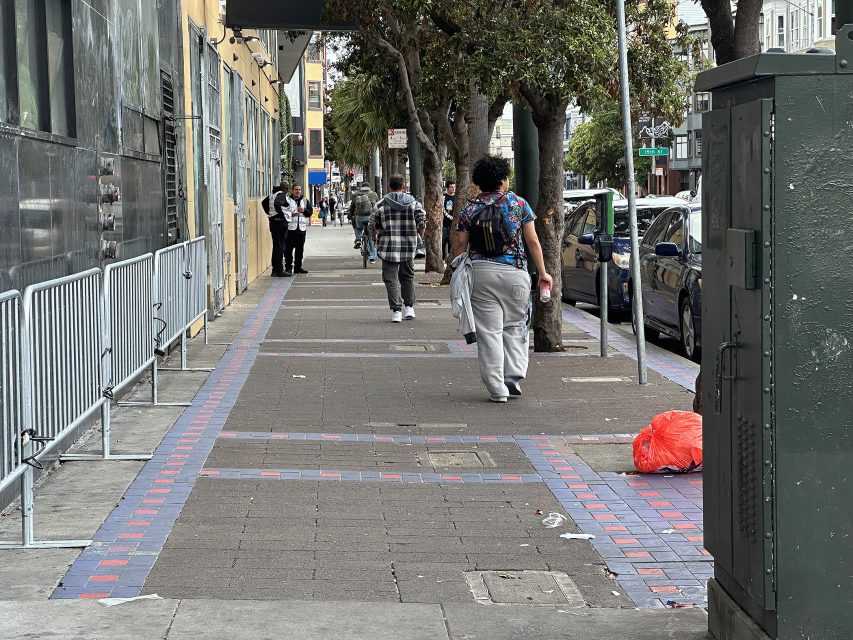 People walk along a city sidewalk lined with trees and parked cars; a red trash bag lies on the ground near a utility box.