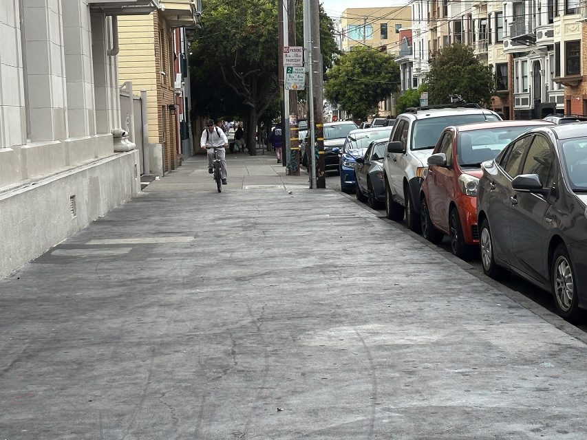 A person rides a bicycle on a city sidewalk beside parked cars and apartment buildings on an overcast day.