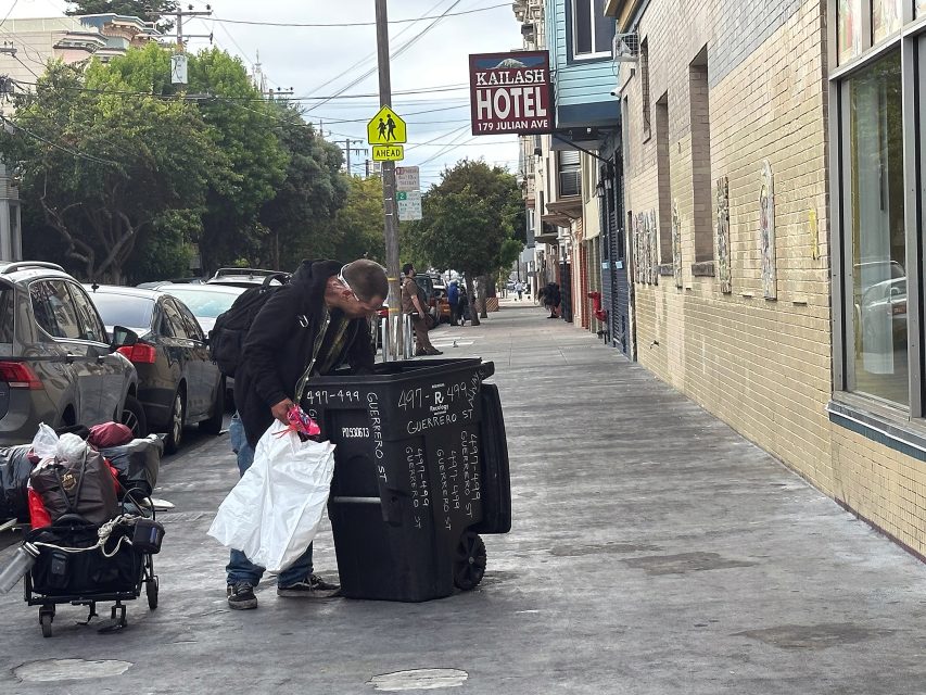 A man searches through a trash bin on a city sidewalk next to parked cars, with a cart and bags beside him. The Kailash Hotel sign is visible in the background.