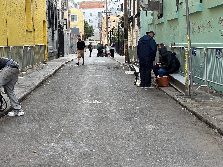 People are gathered and sitting on benches along a graffiti-marked alleyway, with fencing and buildings on either side. A few individuals walk or stand further down the street.
