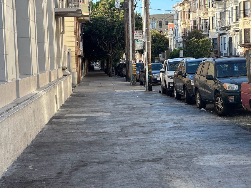 A city sidewalk alongside parked cars and residential buildings on a sunny day. Trees line the street in the background.