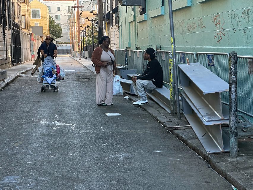 A woman stands and talks to a man sitting on a metal bench in an alley, while another person pushes a stroller and a young child walks ahead.