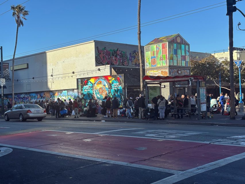 A group of people gather on a city sidewalk near colorful murals and street art, with a small structure and cars parked nearby.