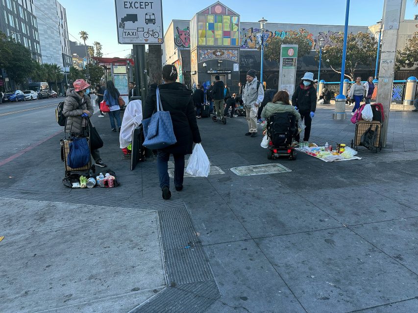 People gather on a city sidewalk, some with bags or in wheelchairs, near street art and a taxi sign; various belongings and items are on the ground.
