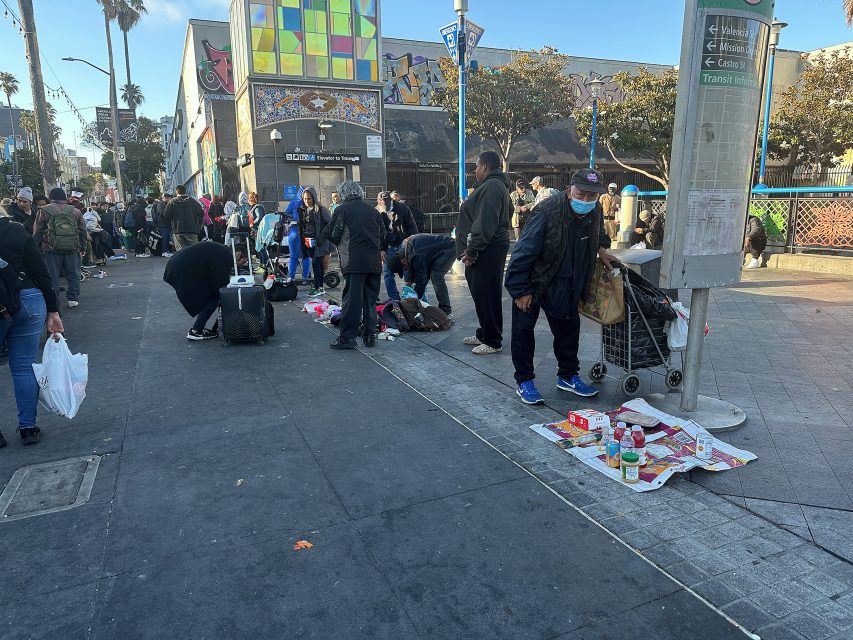 A group of people gather on a city sidewalk with makeshift stalls, personal belongings, and various items laid out on the ground near a transit sign.