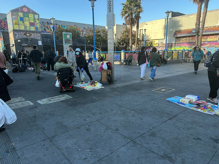 People gather near outdoor street vendors selling goods on the sidewalk in an urban area with graffiti-covered walls and palm trees.