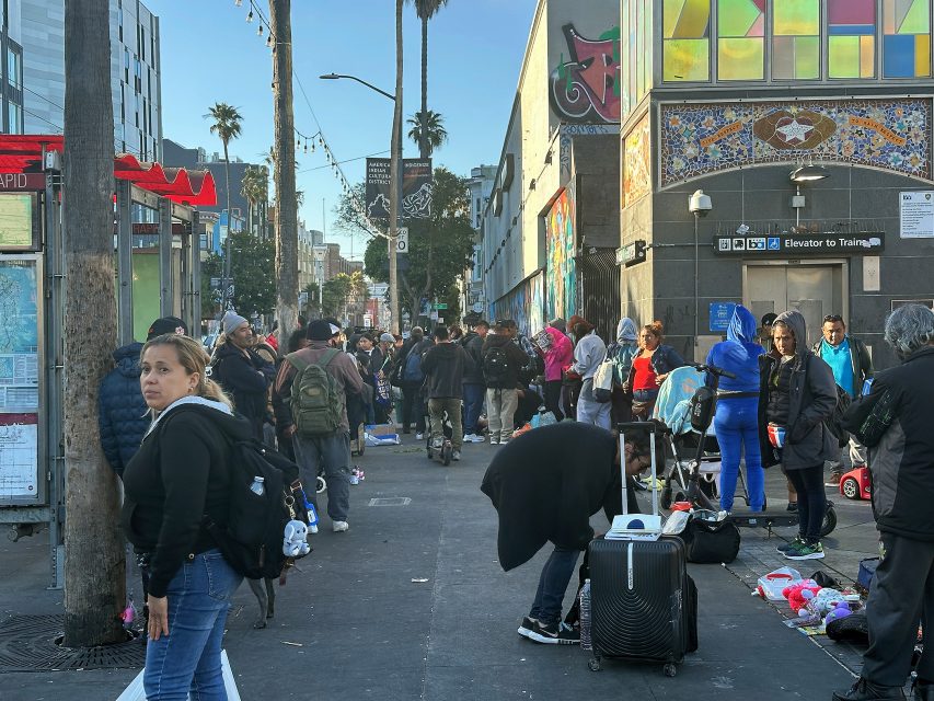A busy urban sidewalk scene with many people, some carrying bags and suitcases, gathered near buildings and street art on a sunny day.