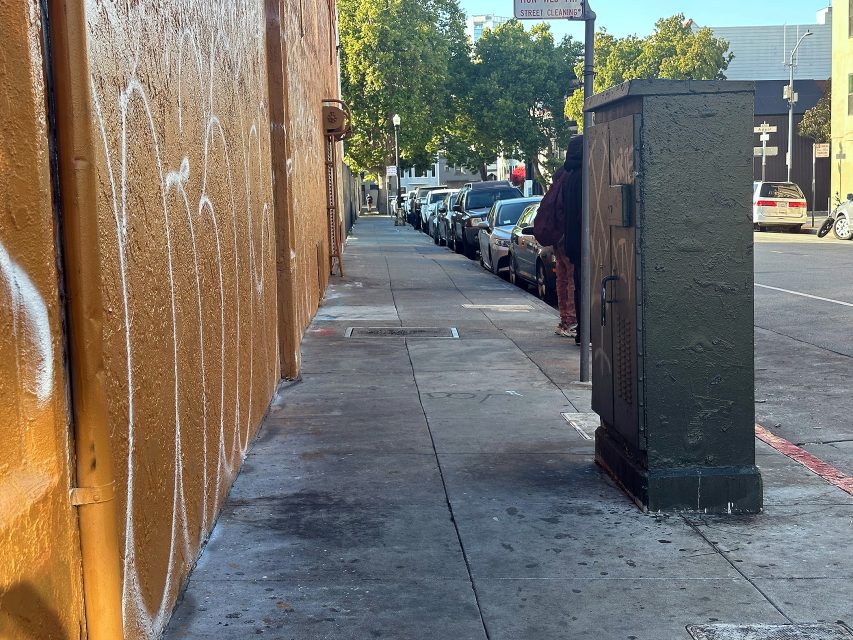 A city sidewalk with a graffiti-covered wall on the left, a utility box on the right, and a row of parked cars along the street in the background.
