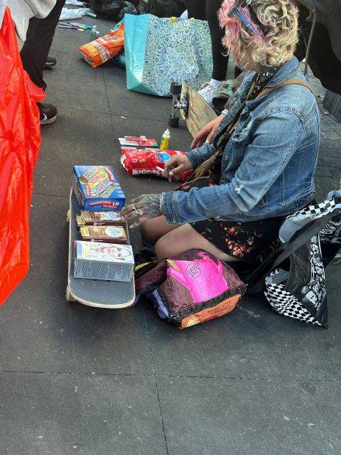 A person with dyed hair in a denim jacket sits on the ground next to a skateboard, sorting packaged food items and bags on a city sidewalk.