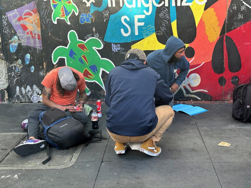 Three people sit and crouch on a sidewalk in front of a colorful mural, with backpacks and belongings scattered around them.
