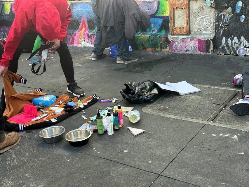 A person in a red jacket bends over a collection of toiletries and supplies laid out on a sidewalk, with graffiti-covered walls in the background.