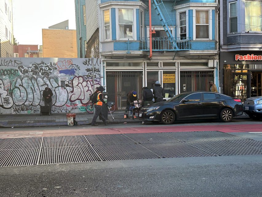 Several people stand and sit near a graffiti-covered wall and storefronts on a city street, with a parked car and a red curb in front.