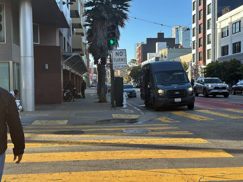 A black van waits at a crosswalk next to a "No Turns" sign on an urban street with yellow pedestrian lines and buildings in the background.