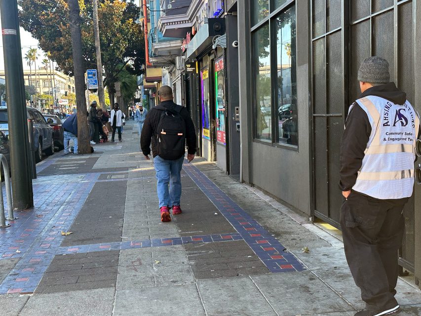 A person in a vest stands by a building while another person with a backpack walks down a city sidewalk; several people are further ahead near storefronts.