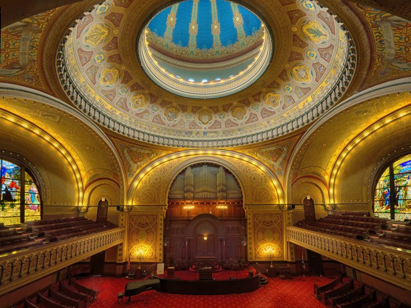 A grand interior of a historic synagogue featuring ornate gold and red detailing, stained glass windows, a large pipe organ, and a domed blue ceiling.