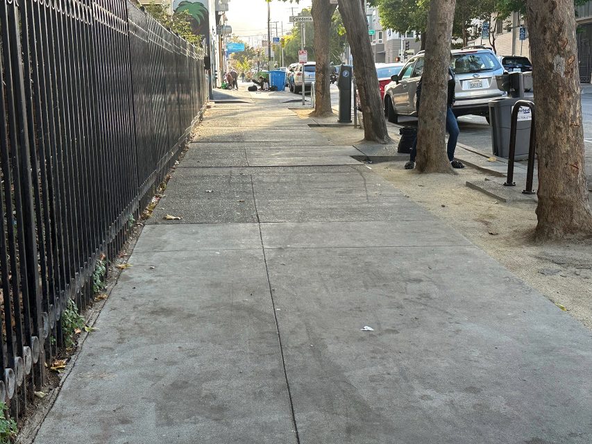 A city sidewalk bordered by a black fence on the left and trees on the right, with parked cars and trash bins visible in the background.