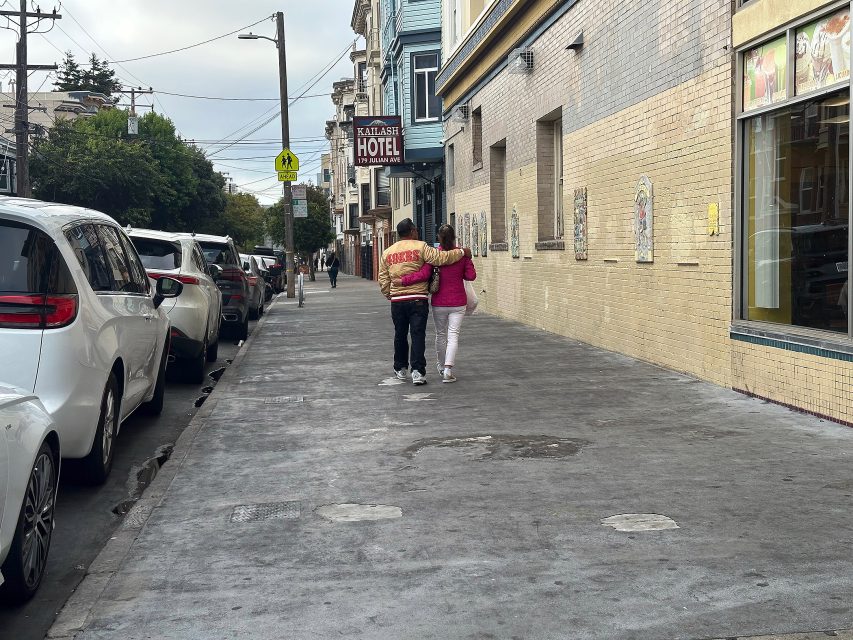 Two people walk with their arms around each other on a city sidewalk lined with parked cars, near a building with a sign that reads "HOTEL.