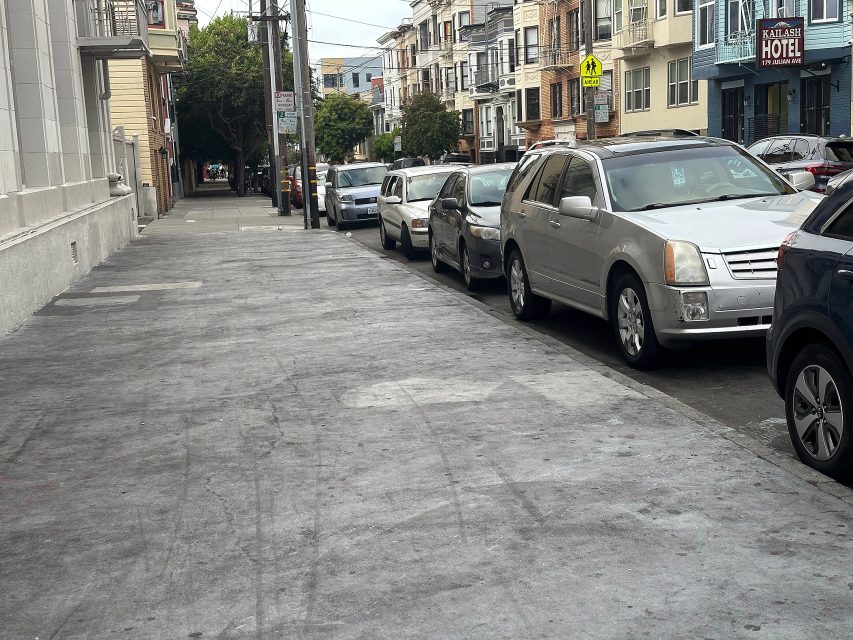 Several parked cars line a quiet city street bordered by apartment buildings, with a hotel and crosswalk sign visible in the background.