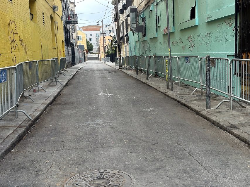Empty urban alleyway with metal barricades on both sides, graffiti on green and yellow walls, and utility wires overhead.