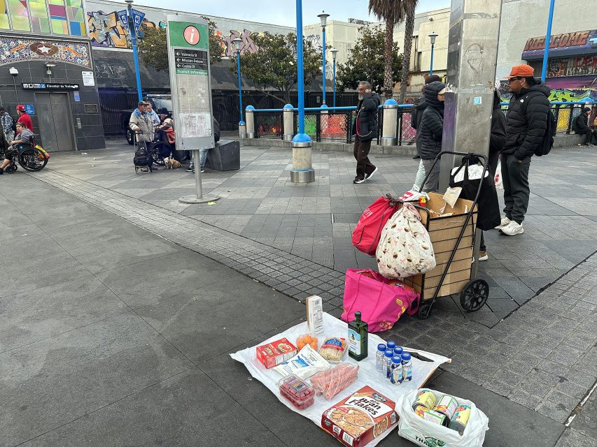 A variety of food items and drinks are laid out on the ground near a market cart, with people standing and walking in a busy urban plaza.