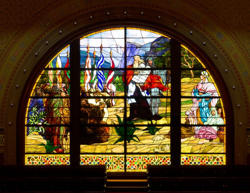 A stained glass window depicting a biblical scene with multiple figures, colorful robes, and a natural outdoor setting, framed by an arched window inside a building.